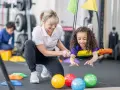 Ergotherapie bei Kleinkindern: Ein Mädchen lacht auf einer Schaukel, während eine Therapeutin sie unterstützt. Bunte Bälle liegen davor.