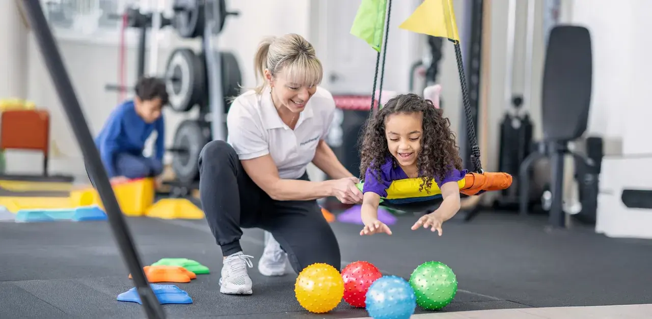 Ergotherapie bei Kleinkindern: Ein Mädchen lacht auf einer Schaukel, während eine Therapeutin sie unterstützt. Bunte Bälle liegen davor.