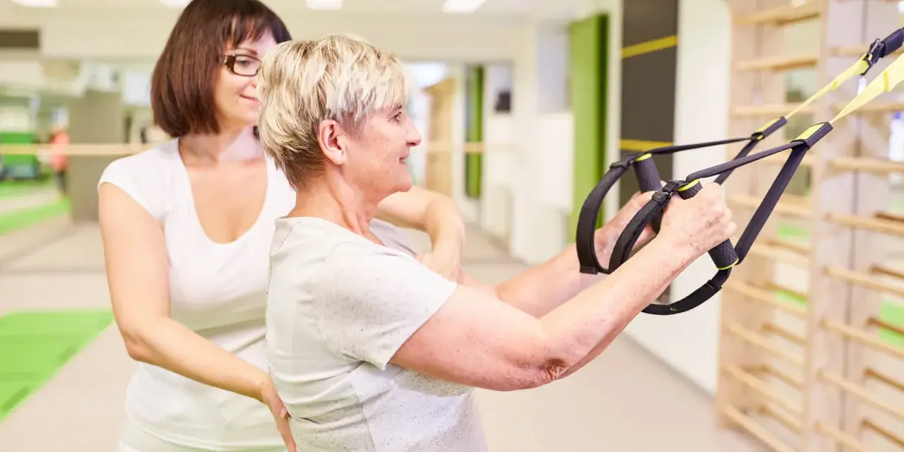 Physiotherapie bei ALS: Eine ältere Frau trainiert mit einem Therapeuten und Suspension Trainer für mehr Kraft und Mobilität.