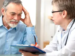 Arzt im Gespräch mit älterem Mann, der sich an die Stirn fasst.