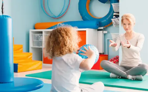 Ergotherapie für Kinder: Eine Therapeutin leitet ein Kind mit lockigem Haar an, das einen blauen Ball hält, in einem bunten Raum voller Spielgeräte.