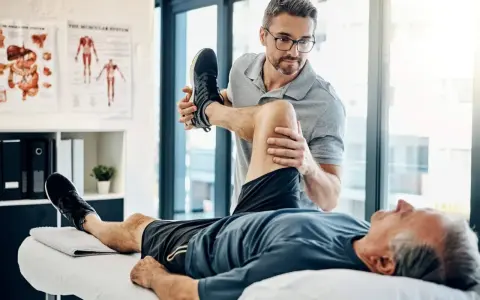 Ein Physiotherapeut dehnt das Bein eines Patienten. Der Patient liegt auf einer Liege, während der Therapeut sein Knie beugt. Wann Physiotherapie? Hier wird geholfen.