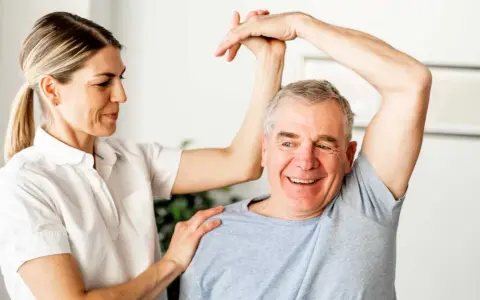 Physiotherapeutin unterstützt Mann bei Übungen zur Wiederherstellung der Beweglichkeit nach Schlaganfall. Verordnung Physiotherapie nach Schlaganfall wird umgesetzt.