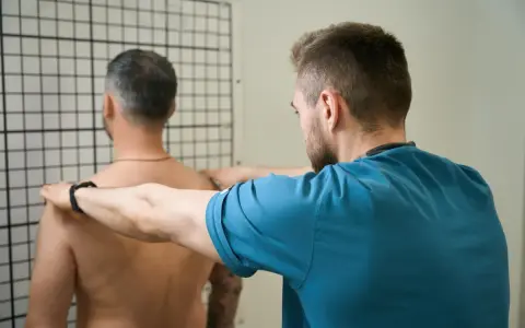 Therapeut führt krankengymnastik schulter Übungen durch. Ein Mann steht vor einem Gitter, während ein Therapeut seine Schultern berührt.