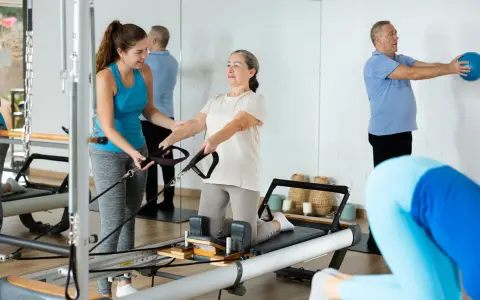 Frauen und Männer trainieren im **Reha-Sport** auf Pilatesgeräten. Eine Trainerin hilft einer älteren Frau, während ein Mann mit einem Ball übt.