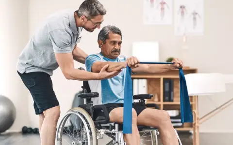 Ein Mann im Rollstuhl trainiert mit einem Therapeuten, der ihm hilft, die Widerstandsbandübungen auszuführen. Die Physiotherapeuten unterstützen ihn bei seiner Genesung.