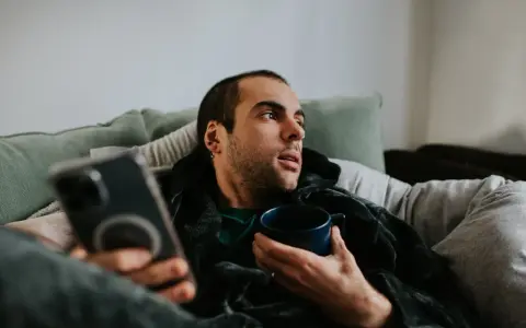 Mann liegt auf Sofa, hält Tasse und Handy. Mögliche Bewegungsmangel Symptome wie Müdigkeit und Lethargie werden angedeutet.