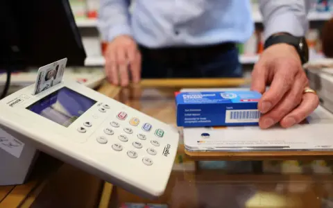 Krankenkassenkarte steckt in Terminal, Hand greift nach Medikamentenpackung.