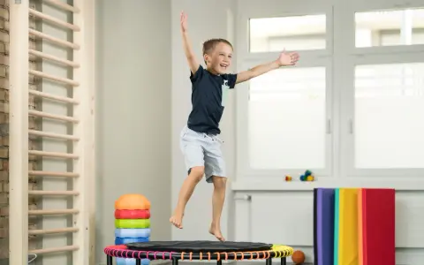 Fröhlicher Junge springt auf Trampolin, Teil der kinder physiotherapie. Kletterwand und bunte Matten im Hintergrund.