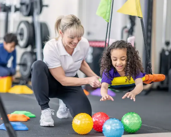 Ergotherapie bei Kleinkindern: Ein Mädchen lacht auf einer Schaukel, während eine Therapeutin sie unterstützt. Bunte Bälle liegen davor.
