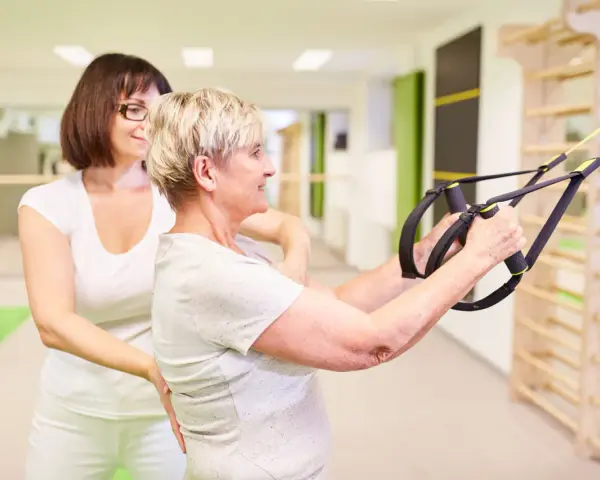 Physiotherapie bei ALS: Eine ältere Frau trainiert mit einem Therapeuten und Suspension Trainer für mehr Kraft und Mobilität.