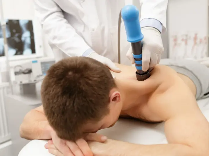 Arzt behandelt HWS-Syndrom mit Stoßwellentherapie. Ein Mann liegt auf dem Bauch, während ein Arzt mit einem blauen Gerät auf seinen Rücken drückt.