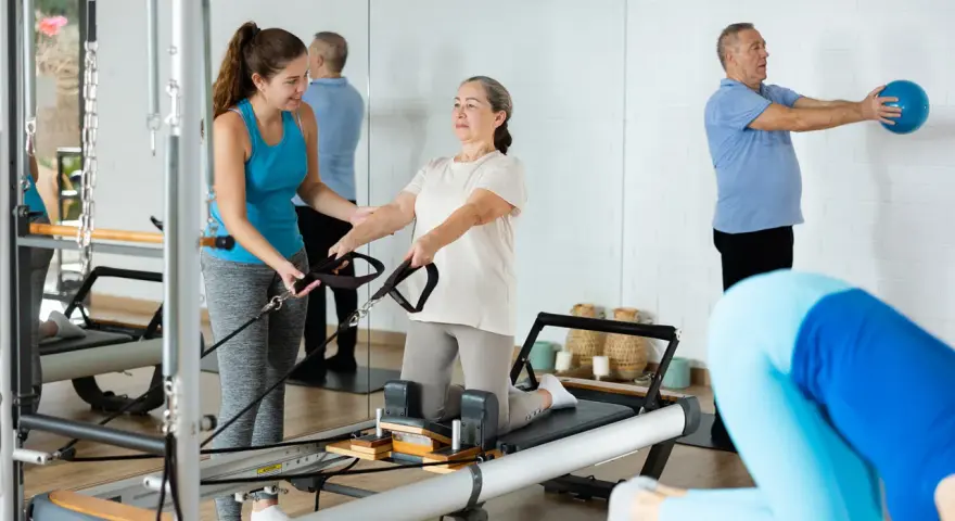 Frauen und Männer trainieren im **Reha-Sport** auf Pilatesgeräten. Eine Trainerin hilft einer älteren Frau, während ein Mann mit einem Ball übt.