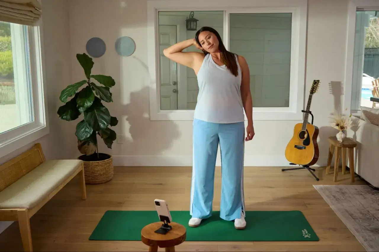 Frau in bequemer Kleidung (Tanktop, weite Hose) übt Nackenübungen zur Physiotherapie. Sie schaut auf ihr Handy.