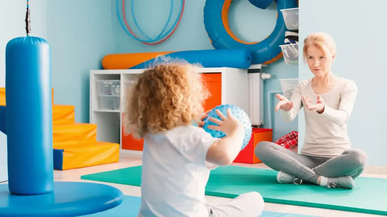 Ergotherapie bei Kleinkindern: Ein Kind mit Locken hält einen blauen Ball, während eine Therapeutin ihm im Lotussitz Anweisungen gibt.