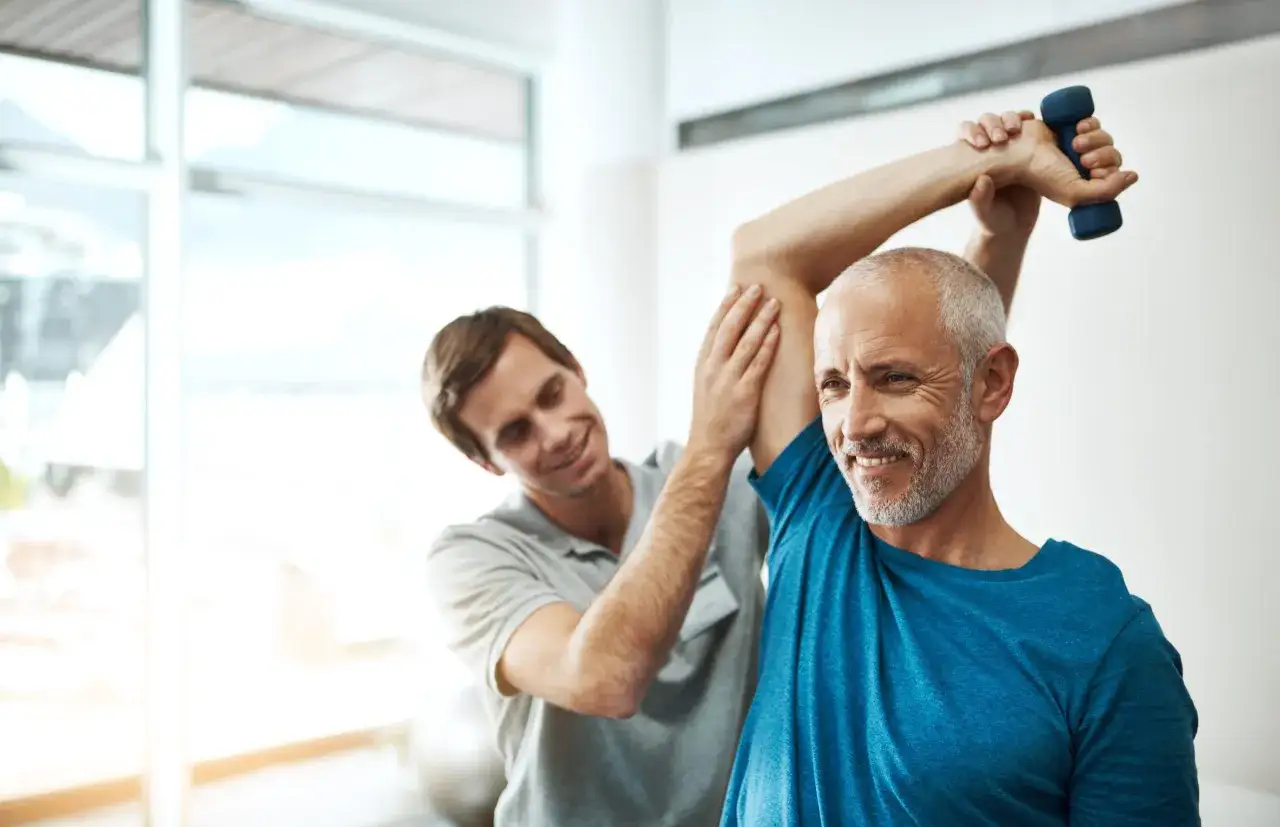 Ein Mann hebt mit einem Physiotherapeuten eine Hantel. Fortschritte nach Physiotherapie sind sichtbar.