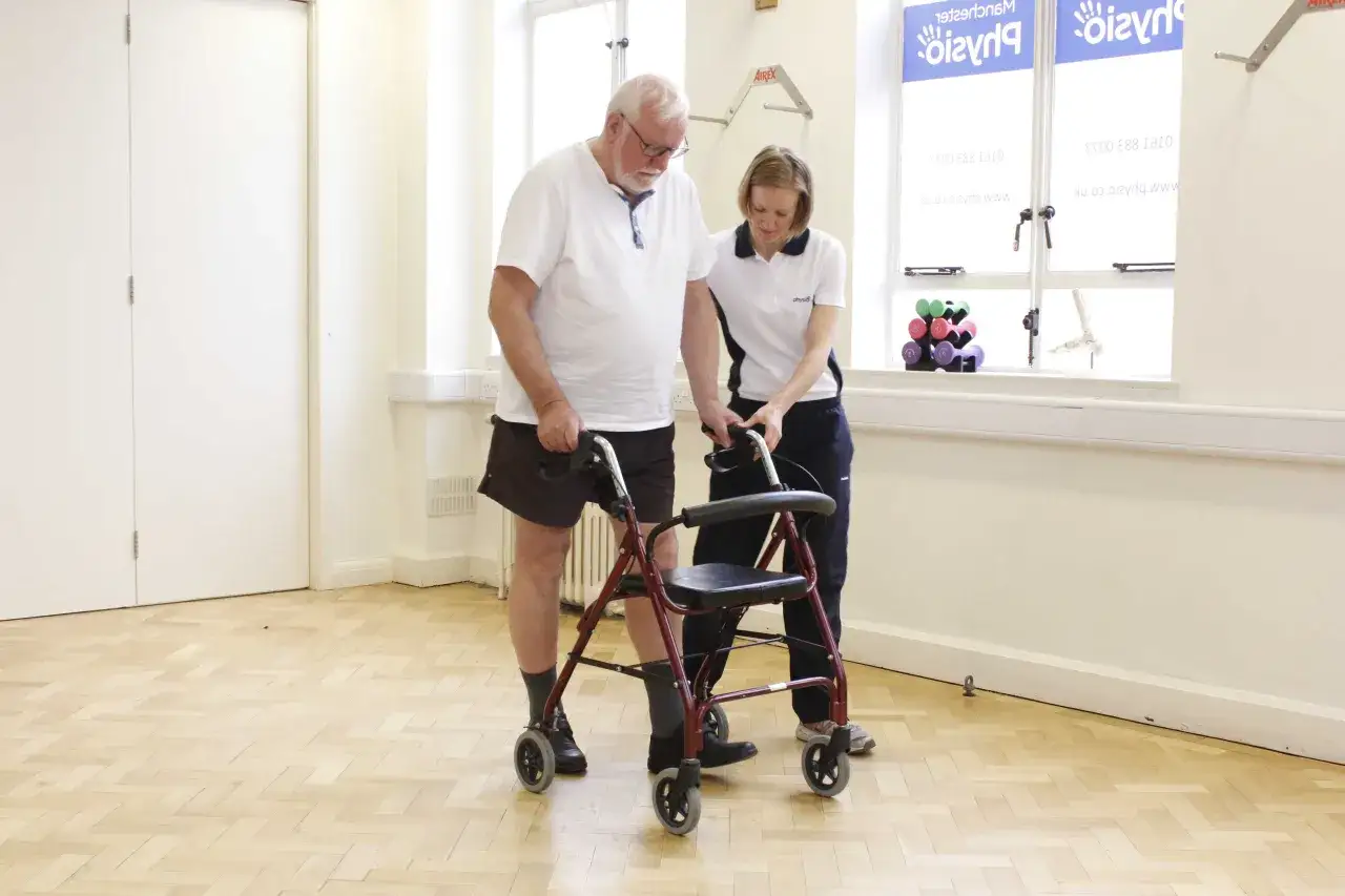 Ein älterer Mann mit Rollator erhält Unterstützung bei der Physiotherapie. Eine Therapeutin hilft ihm beim Gehen.