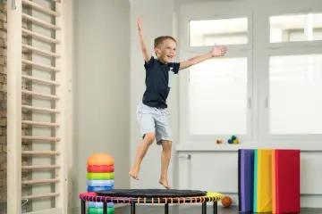 Fröhlicher Junge springt auf Trampolin, Teil der kinder physiotherapie. Kletterwand und bunte Matten im Hintergrund.