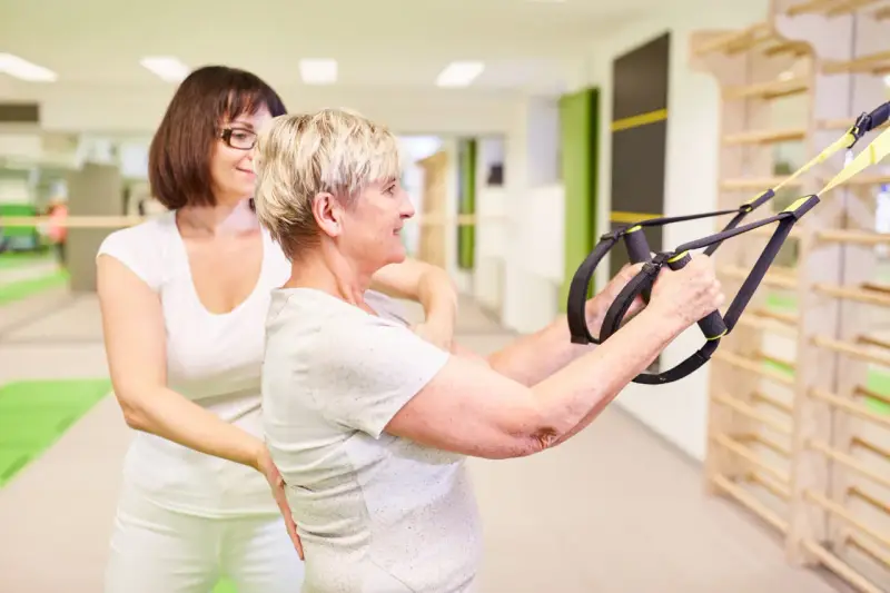Physiotherapie bei ALS: Eine ältere Frau trainiert mit einem Therapeuten und Suspension Trainer für mehr Kraft und Mobilität.